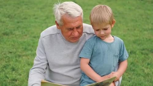 Happy Grandfather Reading Book to Curious Grandson Outdoors Close Up Grandpa with Grandchild