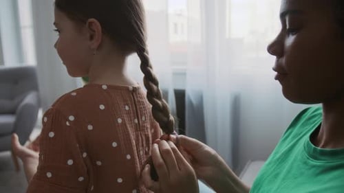 Young Nanny Braiding Hair of Little Girl at Home