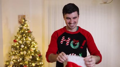 Man wearing festive jumper in front of Christmas tree thrown Santa hat and blows snowball end off hi