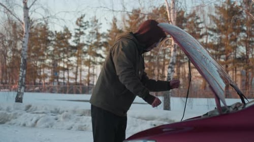 Man in Hoodie Inspects Engine Under Open Bonnet in Winter Street Scene