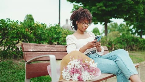 Woman sits in the park on a bench with a flowers with a smartphone in her hands