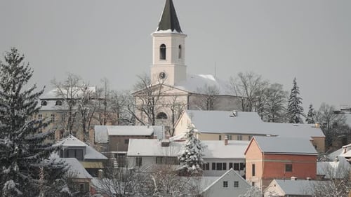 Snow-covered trees and houses create a serene winter view in a quiet Prague neighborhood. A church i