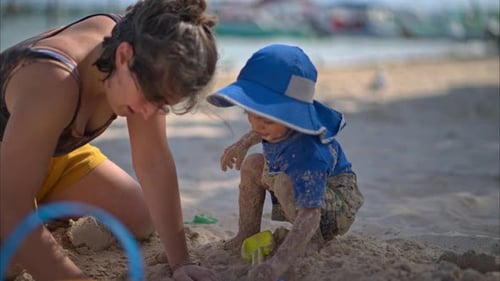 Cute little boy covered in sand playing with his auntie on the beach with toys on a sunny afternoon