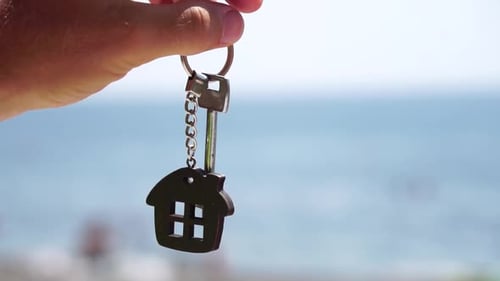 Keys with a Houseshaped Keychain in a Man's Hand on Sea Beach Background