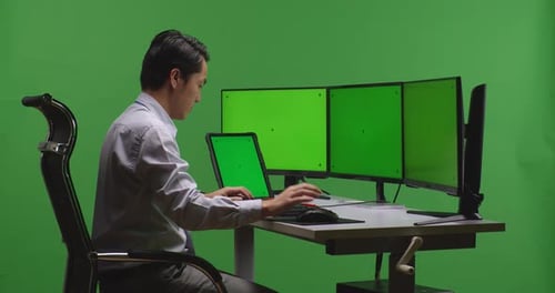 Asiatischer Mann arbeitet mit Tablet und mehreren Monitoren mit grünem Mock-Up-Bildschirm auf grünem Bildschirm