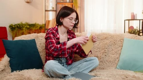 Thoughtful woman opening letter sitting on sofa