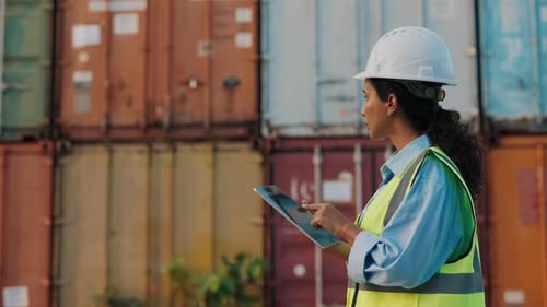 Female Worker Walking Outside With Tablet At Cargo Containers Wearing Safety Uniform