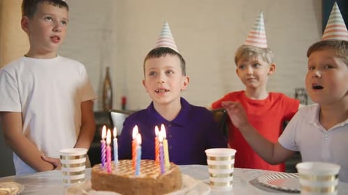 Um aniversariante com um boné festivo apagando as velas de um bolo de aniversário e seus amigos rindo