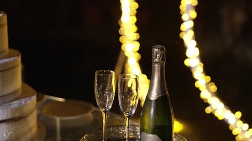 Champagne bottle with two flutes on a table, surrounded by warm fairy lights and a tiered cake in a
