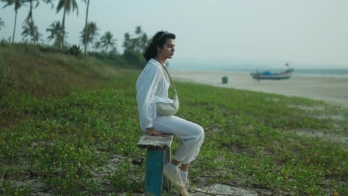 Woman sitting alone on a stool by a tropical beach with palm trees, contemplating the sea, peaceful