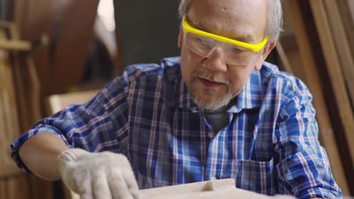 An elderly Asian male carpenter sanding a wooden piece of furniture in a carpentry workshop