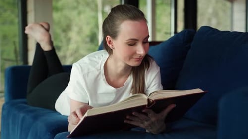 Eine junge lächelnde, fröhliche Frau liest ein Buch, das auf einem blauen Sofa in einem Zimmer liegt
