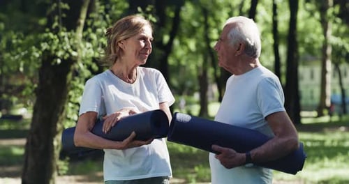 Cinematic shot of smiling senior couple carrying gymnastics mat before start exercising in green p