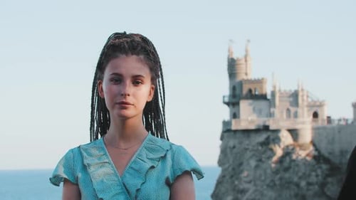 A Young Woman with Dreadlocks Standing on a Background of Sea and a Castle on the Edge Cliff