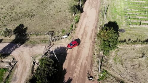 Shot aerial drone footage tracking a red car leaving a house in the country, on an empty road surrou