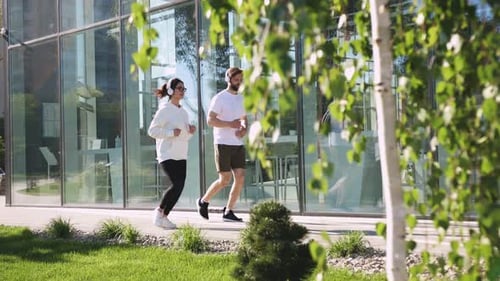 Joyful Caucasian Young Male and Female Joggers in Headphones Jogging Together on Sunny Summer Day