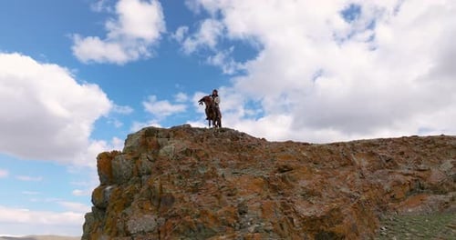 Mongolian Hunter On His Horse Atop Cliff With Eagle Perching On His Arm In Western Mongolia. low ang