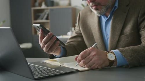 Businessman Using Smartphone and Writing Down in Notebook at Office Desk