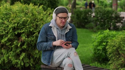 Young Man Enjoying Music While Using Smartphone in a Green Park Surrounded By Trees During a Sunny