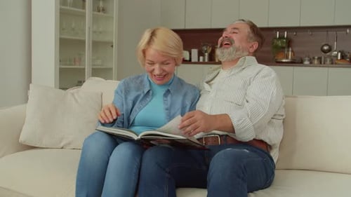 Alegre pareja jubilada viendo el álbum de fotos familiar y compartiendo recuerdos felices