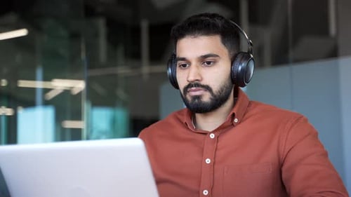 A focused IT specialist in wireless headphones works on a laptop sitting at a workplace in a busines