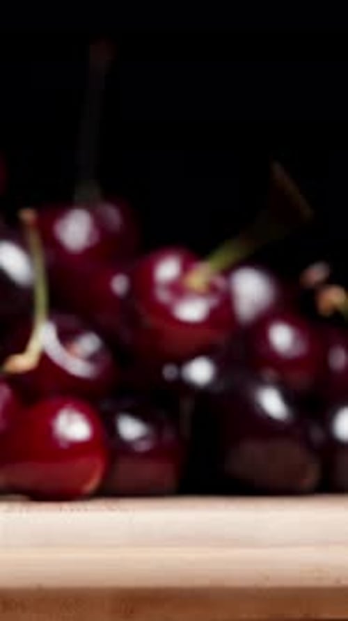 Vertical video. A cherry falls down from a wooden board against a black background, in slow motion.