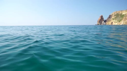 Sea Water Surface Camera Flies Over the Calm Azure Sea with Volcanic Rocky Shores on Background