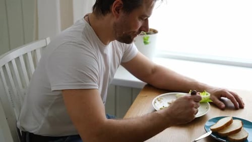 Handsome Bearded Man is Eating Green Fresh Vegan Salad in the Kitchen at Home Male Sitting at Table