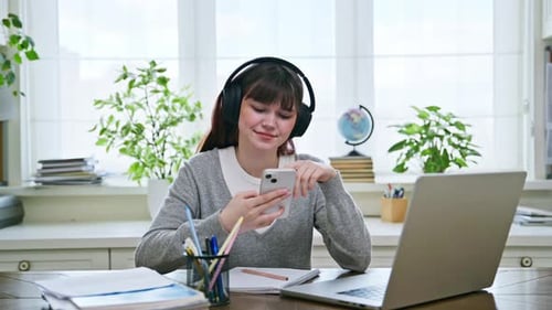 Teenager-Studentin trägt Kopfhörer mit Smartphone und sitzt am Schreibtisch mit Computer