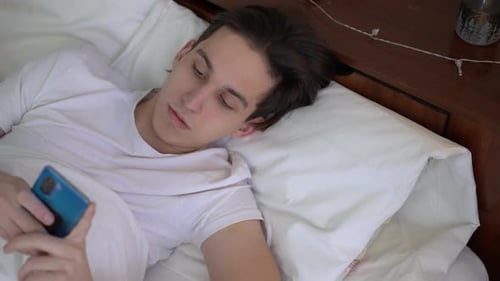 Top View Portrait of Young Man Scrolling Social Media in Phone Application Lying in Soft Comfortable