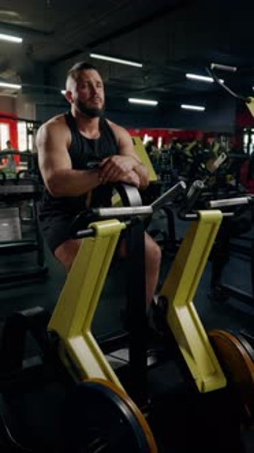 Hombre con barba descansando sobre una máquina de ejercicios en el gimnasio, expresión contemplativa y constitución muscular