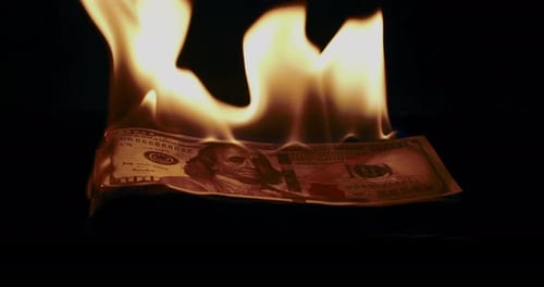 Slow motion low angle wide shot of a stack of prop money as it ignites and burns.