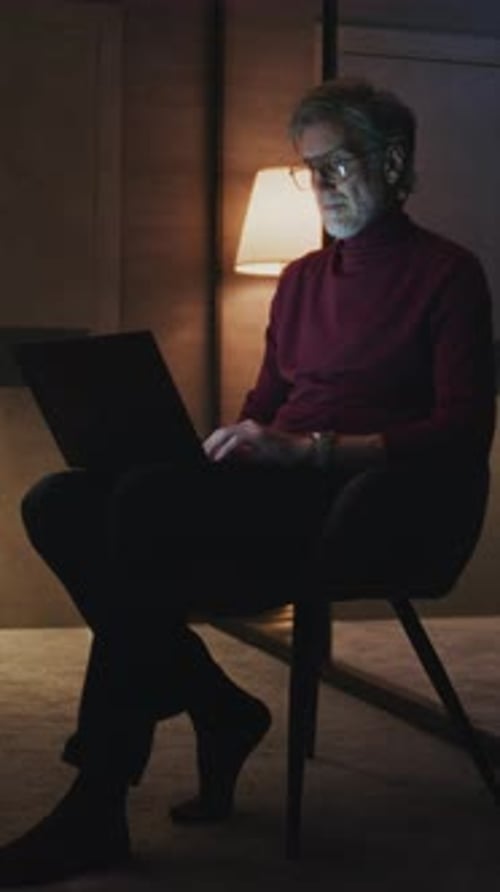 Gray Haired Businessman Wearing Glasses Sits in Modern Hotel Room Typing on Laptop