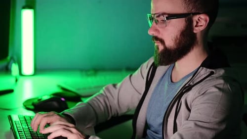 Programmer guy with a beard in glasses works at a computer in a room against the background of a gre