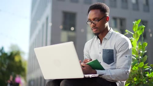 Young african american male student sitting in a city park on a bench with laptop and notebook study