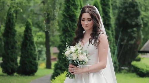 Una hermosa novia se queda sola en el parque con un ramo de novia mira el ramo y luego
