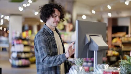 A Guy with Curly Hair and a Mustache in a Plaid Shirt Weighs a Pineapple on a Scale in a Supermarket