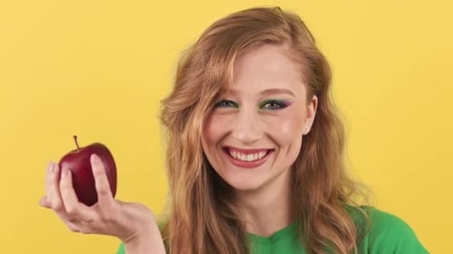 Studio Shot of a Young Redhaired Woman Who Stands on a Yellow Background and Holds a Red Apple in