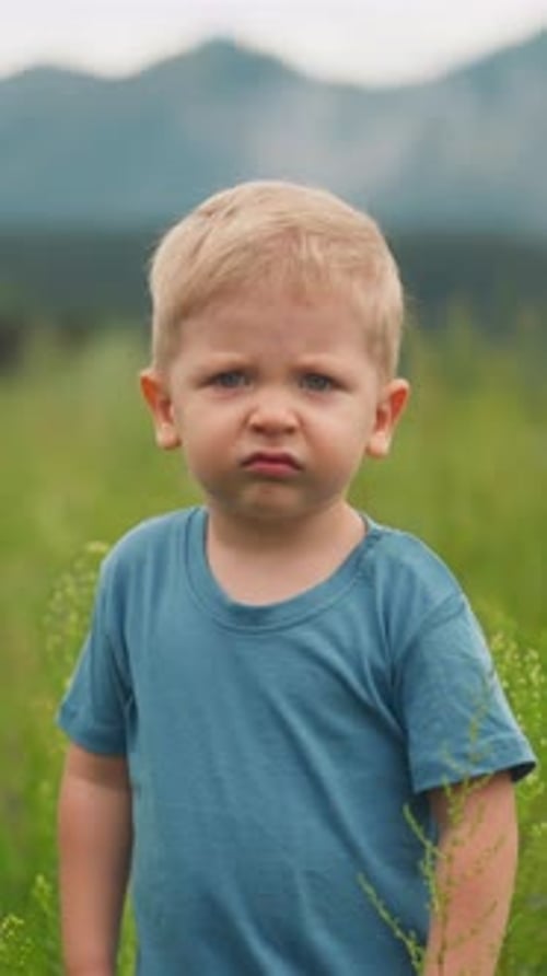 Unhappy Little Boy Grimaces Walking Along Meadow Grass