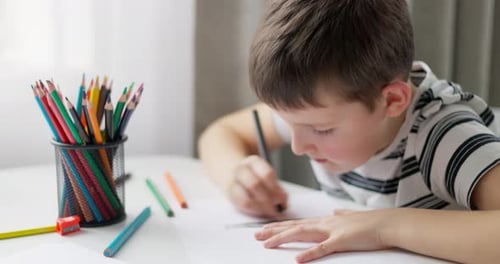 Ein Junge zeichnet mit Buntstiften auf weißem Papier, während er an einem Tisch sitzt