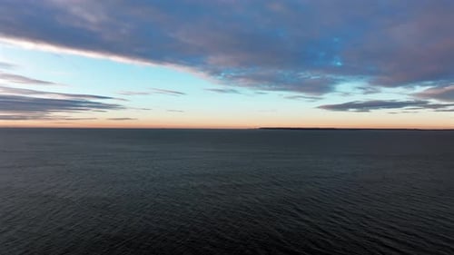 An aerial view over the ocean flying out towards the horizon during sunrise on a cloudy morning. The