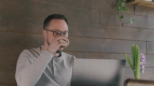Successful Young Businessman with Glasses Using Laptop and Drinks Coffee While Sitting in Cafe in