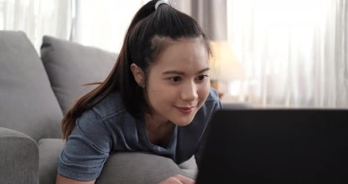 Joven asiática deportiva viendo un Vídeo tutorial de acondicionamiento físico en línea en el portátil mientras se acuesta en el sofá