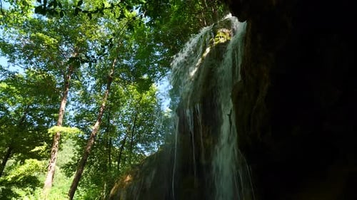 Water falls from the cliff into the cave. Around is green nature.