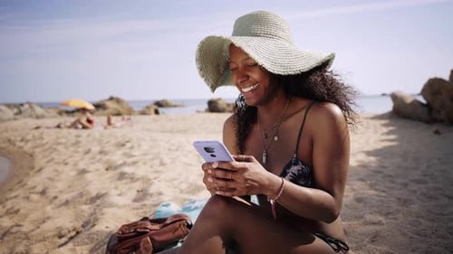 Young Tourist Woman Sitting on the Beach Looking at Her Cell Phone Typing and Laughing
