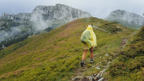 Woman walking on the edge of the mountain wearing a raincoat down looking at foggy forest.
