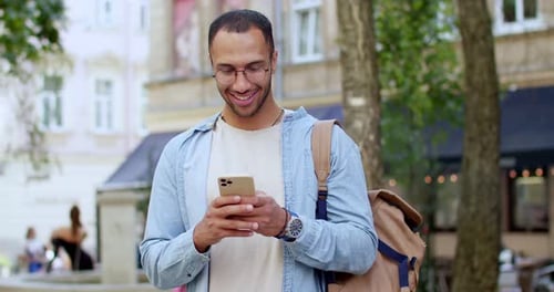 Portrait of Cheerful Young Handsome Man in Glasses Standing at Street Texting Message on Smartphone