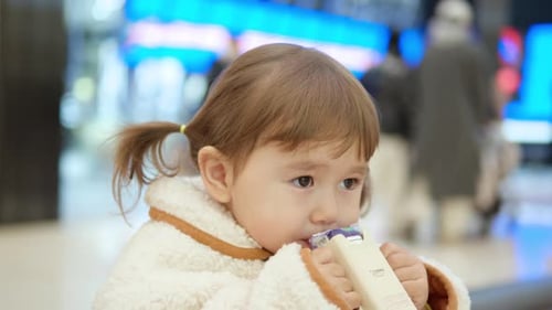 3-year-old Female Child Drinking Juice In The Shopping Mall Cafe, Shoppers Walking By In Blurry Back
