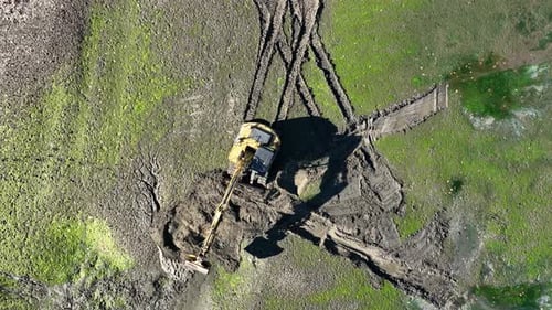 Excavator digging on muddy terrain surrounded by green patches seen from above on a sunny day