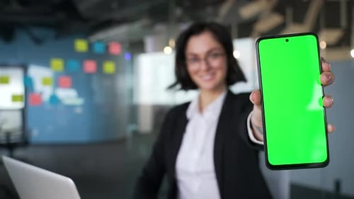 Professional female in modern office environment presenting phone with green screen. Color key,